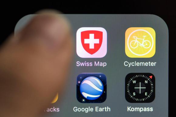 أصبع إبهام يتحرك فوق شاشة جهاز هاتف إيفون تعرض تطبيق الخرائط السويسرية وتطبيق غوغل إيرث.