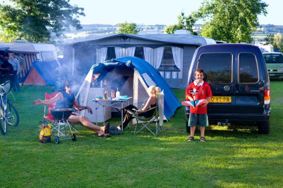Eine Familie sitzt auf dem Campingplatz, die Eltern grillieren und der Sohn spielt mit einem Modellflugzeug.