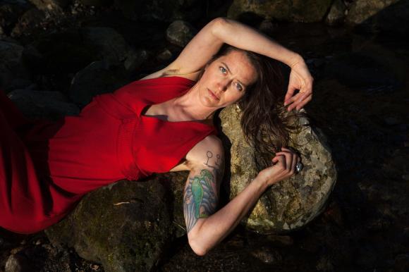 Die Musikerin Bibi Vaplan räkelt sich in einem roten Kleid über einem Felsen. Auf dem linken Arm ist ein Tattoo zu sehen.