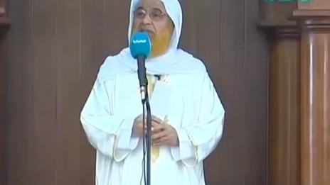 A man in a white robe and orange beard speaks into a microphone