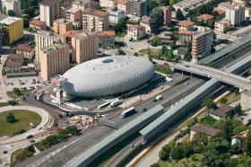 A large lozenge shaped white building sits next to a highway and other buildings