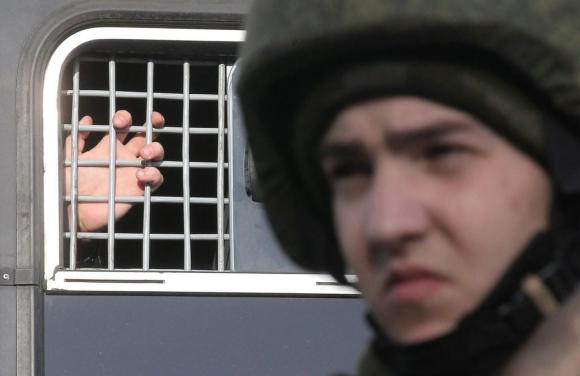 Bild einer Hand, die sich an einem vergitterten Fenster eines Militärfahrzeugs festhält.