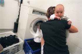Una mujer, con un bebé en los brazos, mete ropa en una lavadora