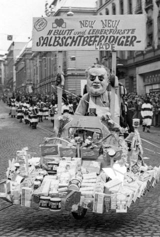 A papier-mâché figure driving a cart filled with consumer products, with a sign above its head with writing on it.