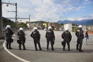 Sei poliziotti in tenuta antisommossa, disposti in rango osservano un piazzale ferroviario