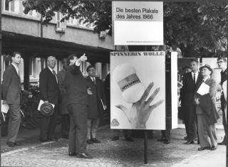 Black and white photo showing a group of men looking at a advertising poster depicting a ball of wool and a hand
