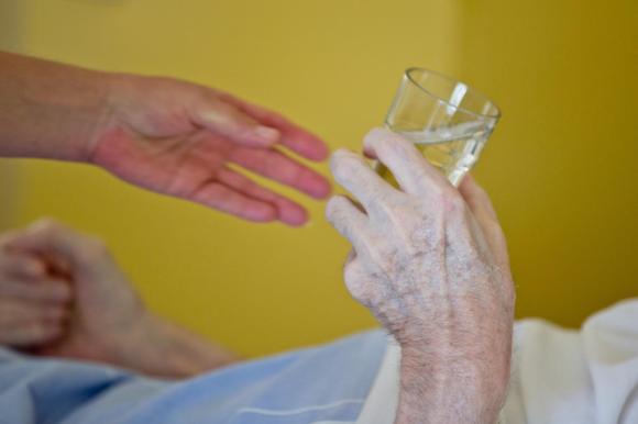 die Hand einer jungen Pflegenden und die Hand einer betagten Person, die ein Glas hält.