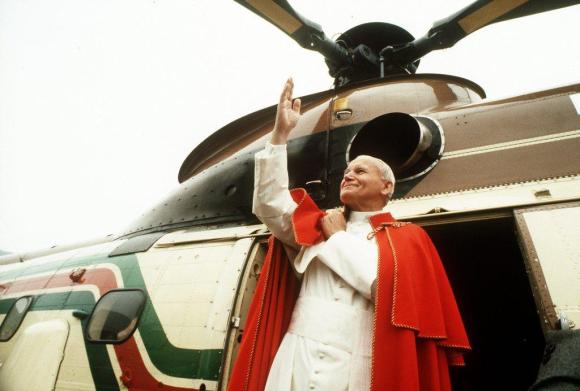 A man wearing a red cape make a sign of greeting while disembarking a helcopter
