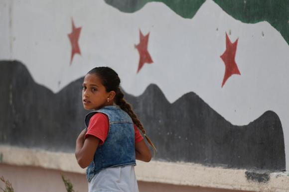 Ein Mädchen geht vor einer mit der syrischen Fahne bemalten Wand und schaut nach links.