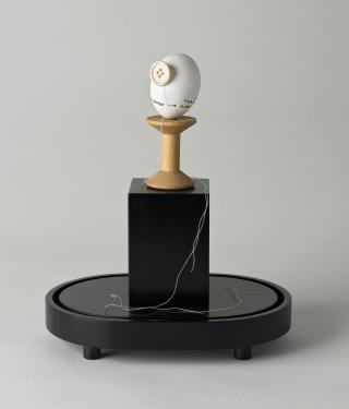 Ein Ei, ein Knopf, eine Fadenspule und Faden auf einem Posdest als Kunstwerk
