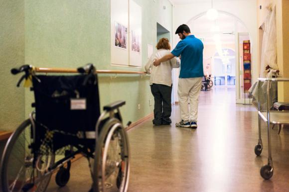 ممر داخل عيادة صحية وفيه كرسي متحرك وممرض يُساعد مريضا على المشي