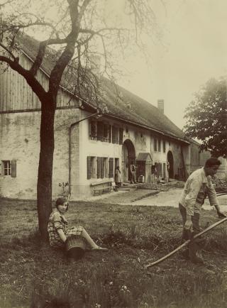 Farmhouse with people in the foreground, woman looking at the camera
