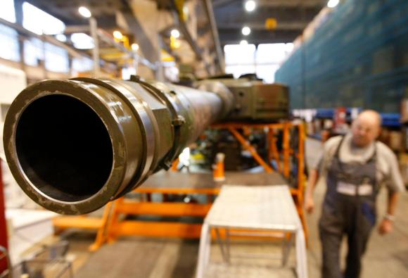 Primo piano di un cannone da carro armato; attorno, sfocato, ambiente industriale e un operaio