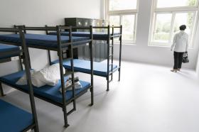 Dormitorio de centro de asilo con dos literas vacías y una persona de pie ante una ventana