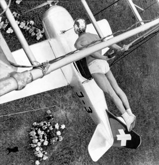 A male acrobat trapezing above an old fashioned aeroplane in mid-flight.