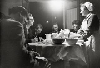 A group of men take notes while a nurse demonstrates childcare with a replica baby.