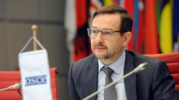 Ein Mann mit Brille sitzt an einem Tisch, auf dem ein Fähnchen mit der Aufschrift OSCE steht.