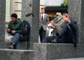 two men sitting on stone bench reading and eating, white collar worker in background