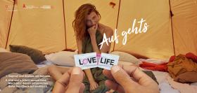 Poster Love Life 2019: unas manos abren un condón. Detrás hay una chica sentada en una cama y semicubierta con una manta