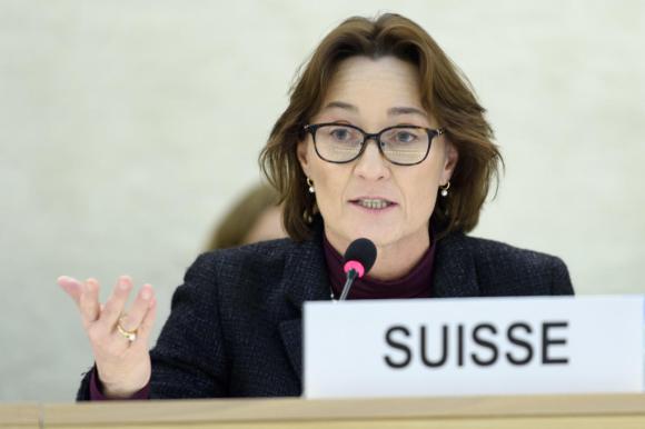 Eine Frau mit Brille sprich in ein Mikrofon, vor ihr auf dem Tisch steht ein Schild mit der Aufschrift Suisse.