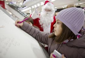 Una niña escribe en una gran hoja de papel. Junto a ella, Papá Noël.