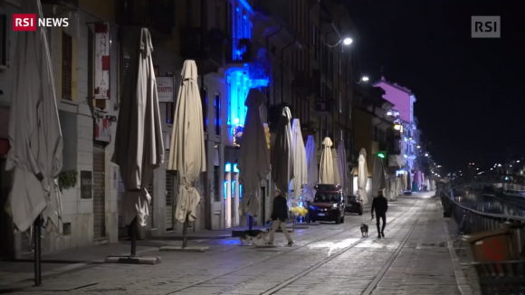 Viale lungo un naviglio con locali e ombrelloni chiusi, poche luci, due uomini portano a spassso il cane