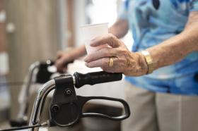 Imagen parcial de una persona mayor empujando un andador y con un vaso en la mano