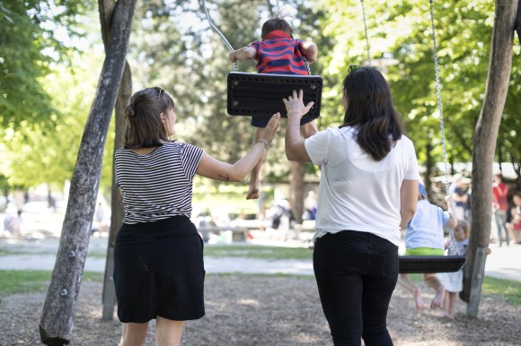 Zwei Frauen stehen zusammen bei einem Kind auf einer Schaukel.