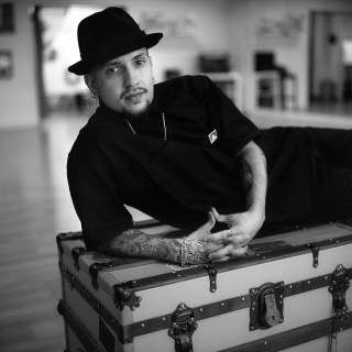 A man with tattoos, wearing a hat, leaning on a box-chest.