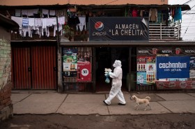 Una persona con mascarilla y vestimenta de protección camina por un barrio pobre con unas cajas de alimentos