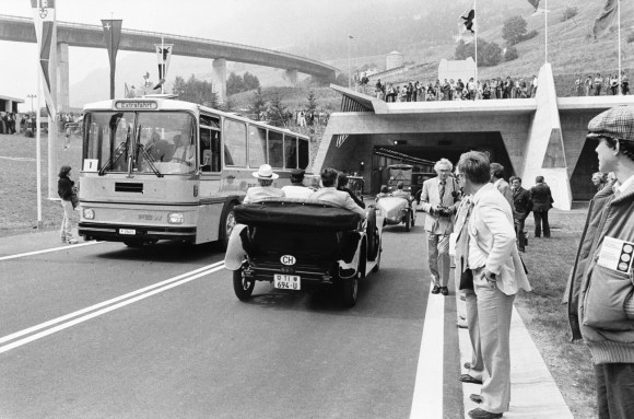 Tunnelportal aus Beton; ein Bus kommt aus dem Tunnel, Oldtimer fahren hinein; Zuschauer am Strassenrand