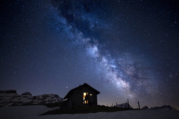 Sternenhimmel bei idealen Lichtverhältnissen in einer alpinen Umgebung; Kleine Hütte mit beleuchtetem Fenster im Vordergrund