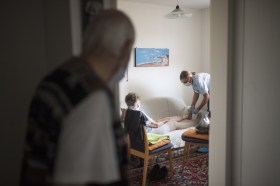 ein alter Mann beobachtet im Wohnzimmer, wie eine Haushaltshilfe sich um seine Frau kümmert
