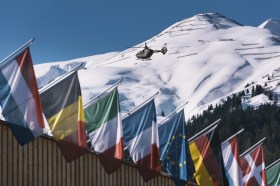 Helicopter flies above flags against mountain backdrop.