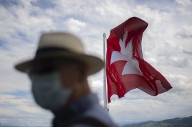 Un homme masqué devant un drapeau suisse.
