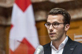 Homme parlant au micro devant un drapeau suisse