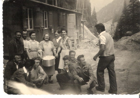 b/n Tre giovani donne e un anziana posano con un pentolone accanto a un gruppo di operai fuori da una baracca