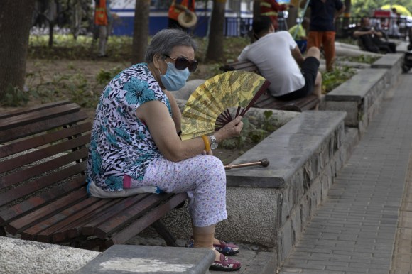 امرأة تجلس فوق مقعد في حديقة وهي ترتدي قناعا وتستخدم مروحة يدوية