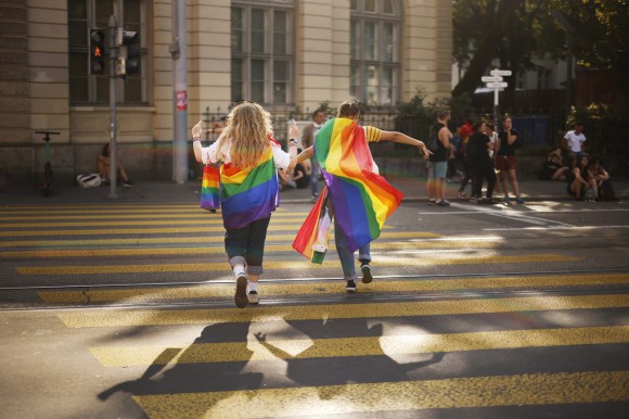 Two women wearing rainbow flags crossing a street