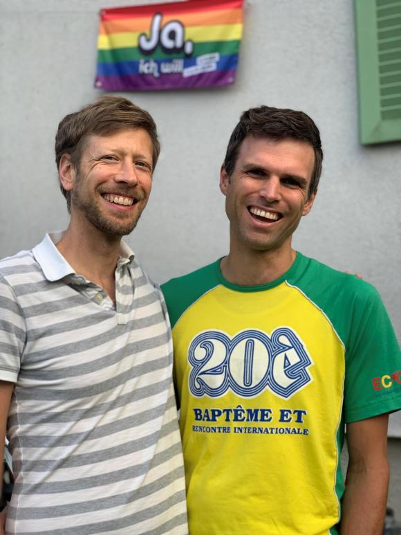 gay couple smiling in front of a rainbow flag
