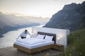 A hotel bed is made on a veranda overlooking a lake and mountains