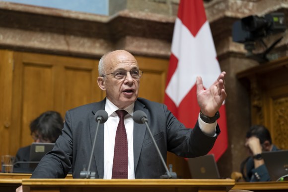 رجل يتحدث أمام مصدح وف يالخلفية علم سويسرا