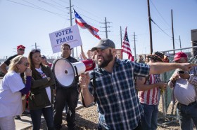 Mann mit Megaphon an einer Demonstration mit USA-Flaggen und einem Schild mit Wahlbetrug.